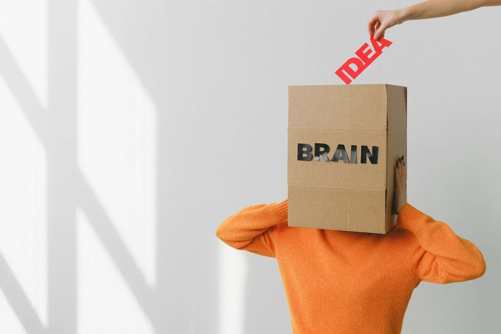 A person wearing an orange sweater holds a cardboard box labeled "BRAIN" over their head while another hand places the word "IDEA" into the box, symbolizing thoughts, creativity, and memory.