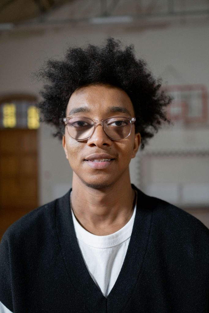 A young man with curly hair and glasses stands in a gymnasium, wearing a black and white sweater. He has a calm and confident expression.