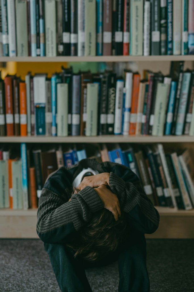 A boy sits on the floor with his head in his lap and arms wrapped around his legs, appearing distressed. He is in a library, surrounded by bookshelves filled with books.