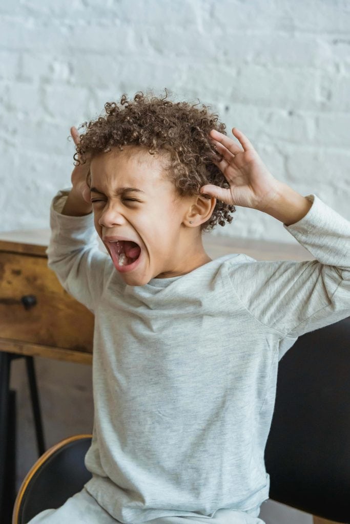 A young boy with curly hair is shouting with his hands on the sides of his head. He appears frustrated or upset, sitting indoors with a white brick wall and a wooden table in the background.