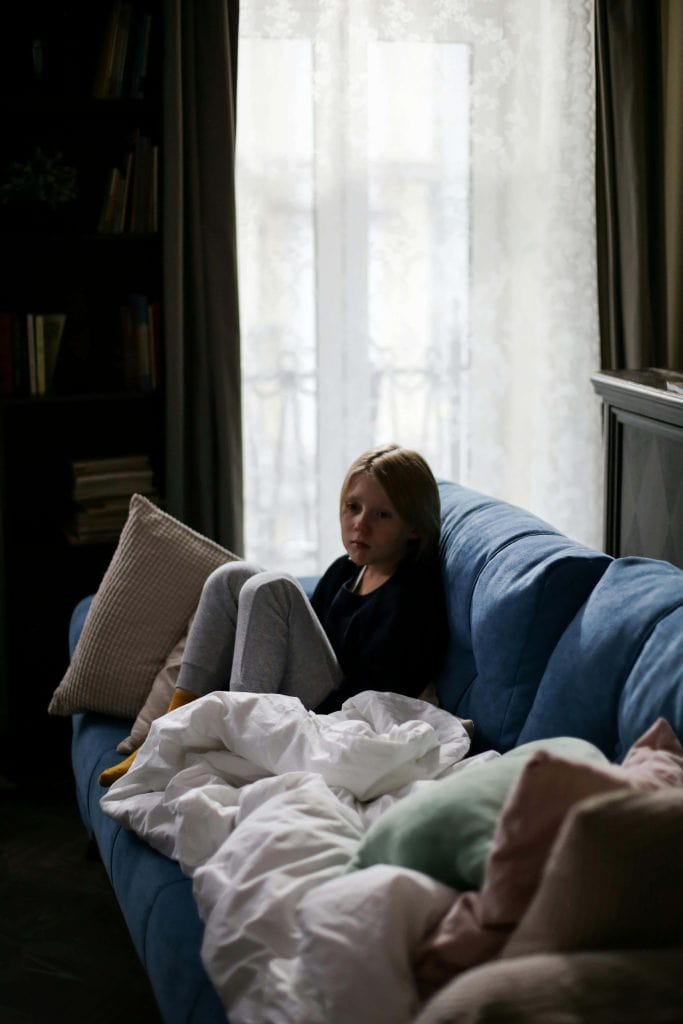 A young girl sits curled up on a blue couch near a window, looking distressed. She is surrounded by pillows and blankets in a dimly lit room with bookshelves in the background.