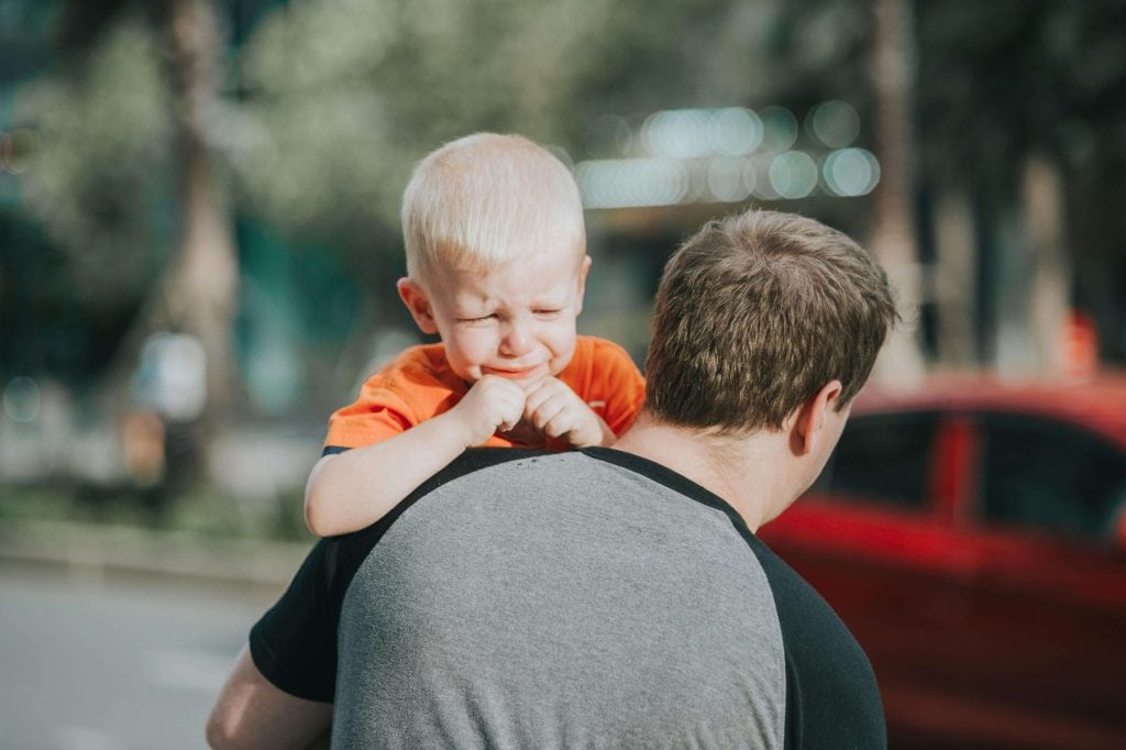 A young child in an orange shirt cries while being carried by a parent, showing signs of frustration. This image represents the challenges of emotion regulation in children.