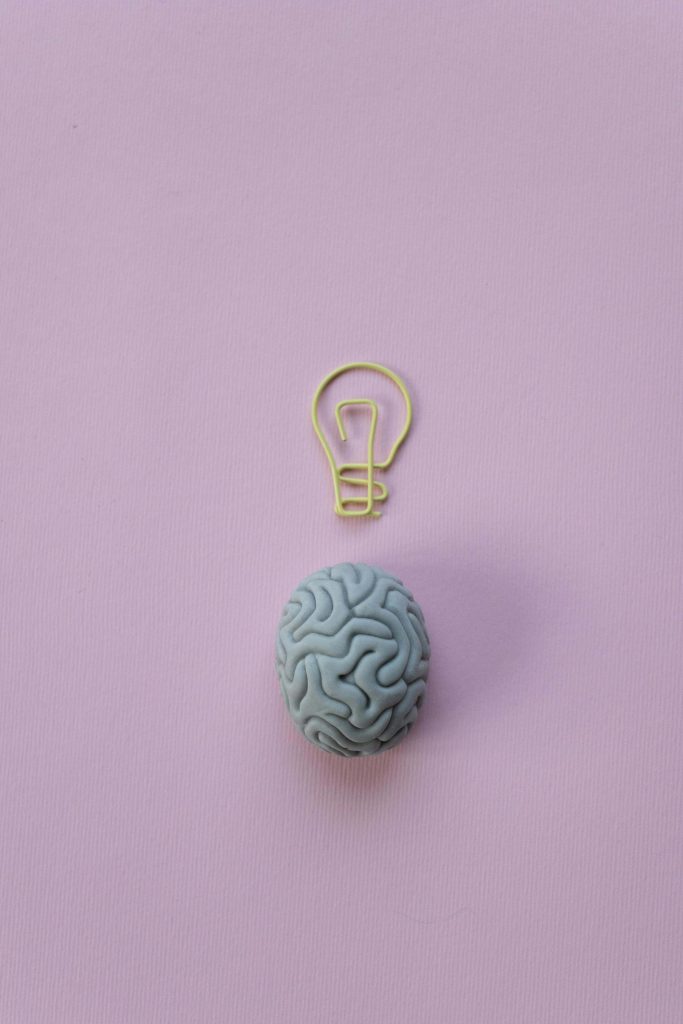 A small blue brain model placed on a pastel pink background with a yellow wire shaped like a lightbulb above it, symbolizing ADHD, creativity, and neurodivergent thinking