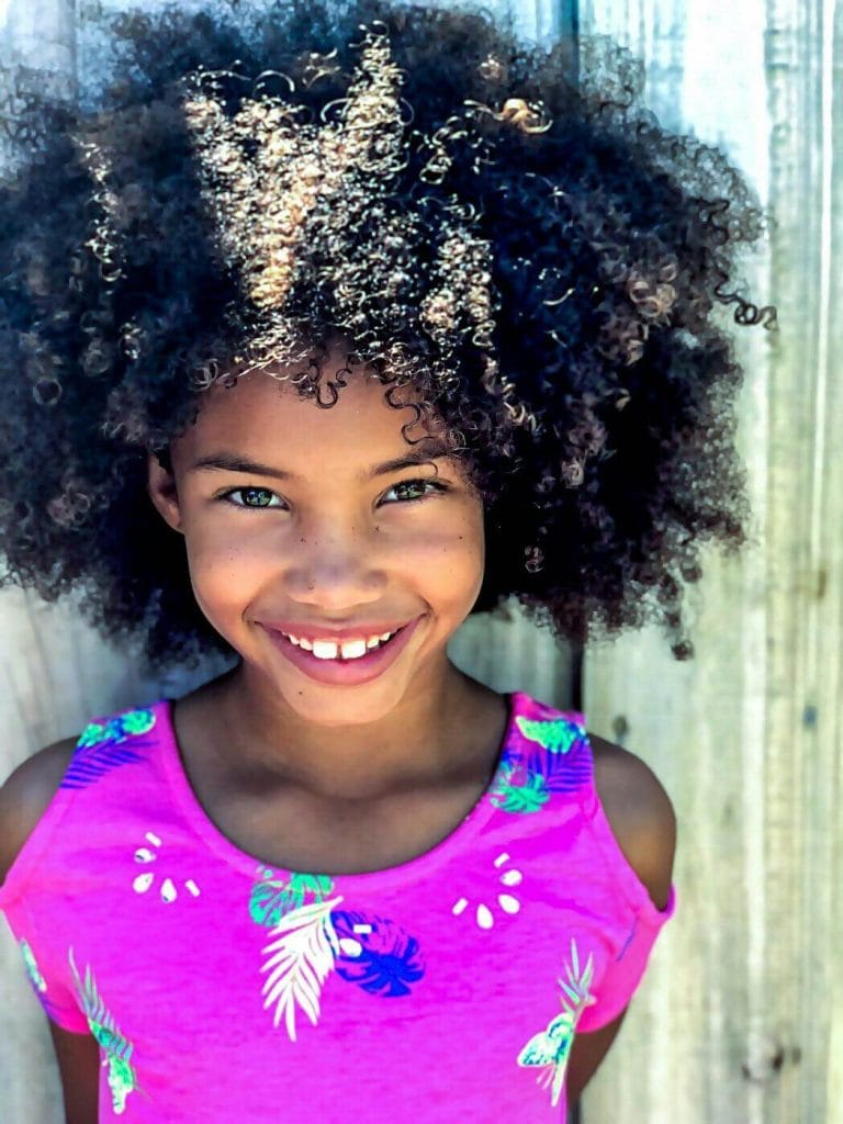 A young Black girl with a bright smile and voluminous curly hair stands in front of a wooden fence, wearing a vibrant pink top with a tropical pattern. Sunlight highlights her natural curls