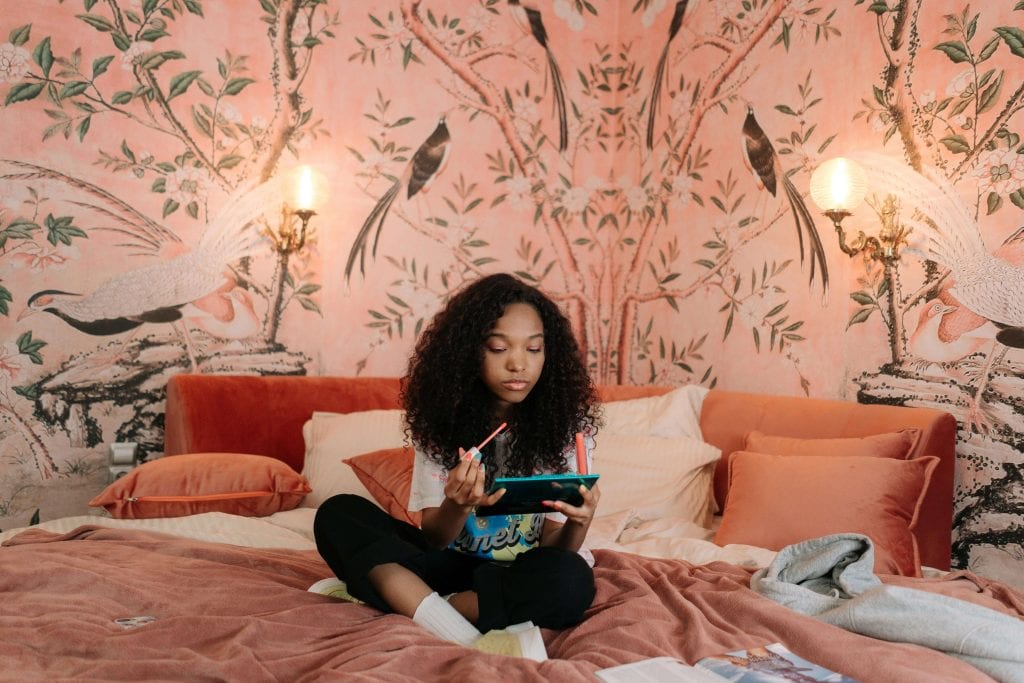 A teenage girl with curly hair sits cross-legged on her bed, using a tablet with a stylus. The room has a warm, pink-toned aesthetic with bird-themed wallpaper and soft lighting