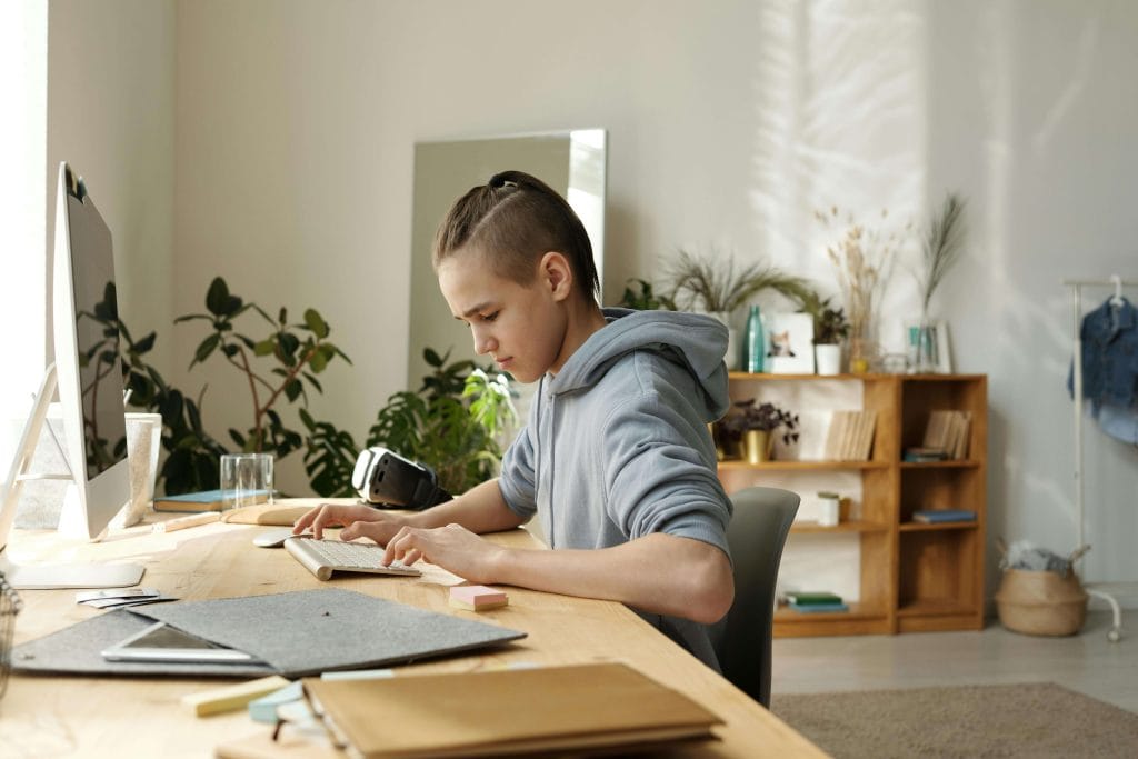 A teenage boy with a half-shaved hairstyle and a hoodie sits at a wooden desk, concentrating on typing. His workspace is organized, with plants and bookshelves in the background