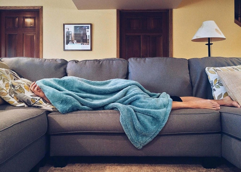 Person resting on a gray sectional couch, covered by a plush blue blanket, showing how weighted blankets can provide comfort in any space.