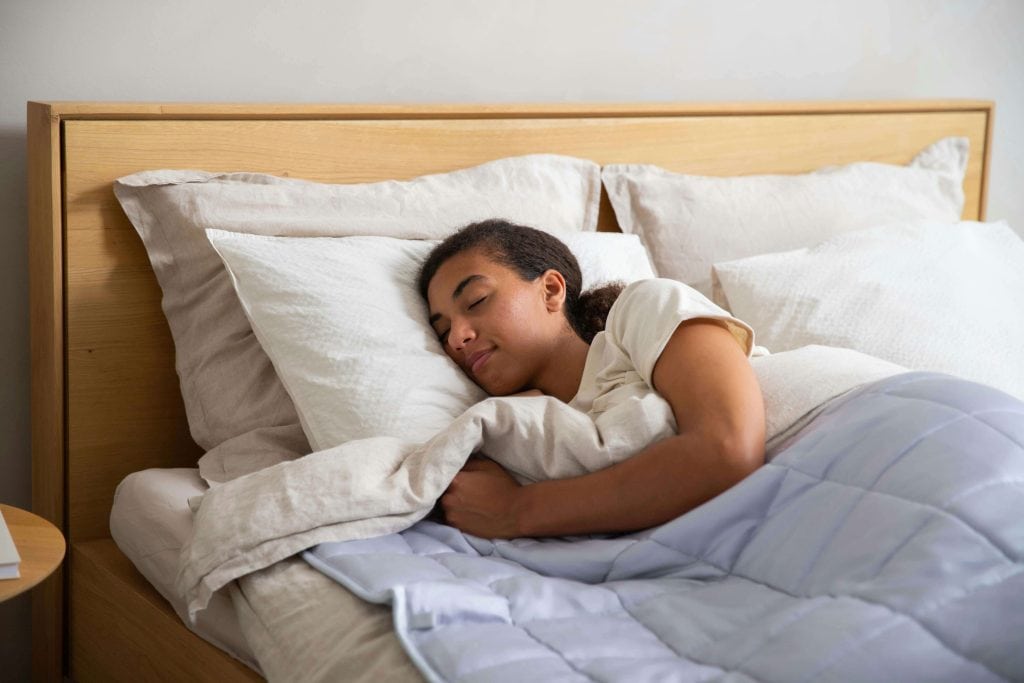 Woman peacefully sleeping on a bed, cuddled under a light blue weighted blanket and surrounded by soft white pillows. weighted blankets for kids with autism