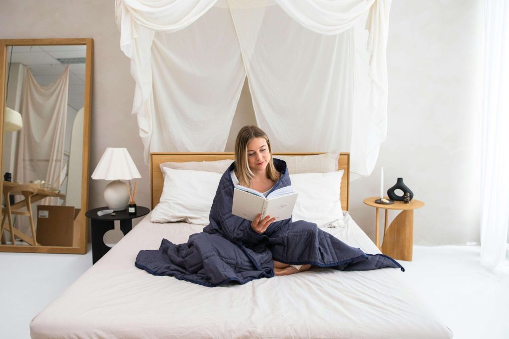 Woman sitting on a bed, wrapped in a navy weighted blanket, reading a book in a serene bedroom with white curtains and minimalist decor.