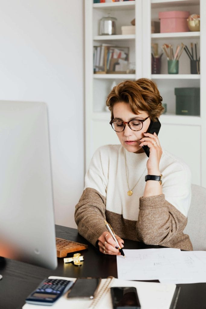 A professional woman on the phone and writing on papers at her desk, representing specialists or educators developing AAC communication plans for children with autism.