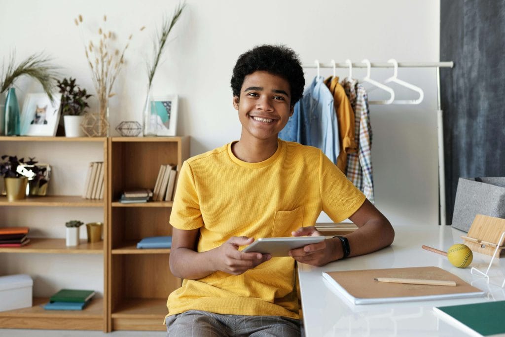 A teenage boy smiling while holding a tablet, sitting at a desk in a home learning space, representing the use of AAC devices for independent communication in autistic teens. aac devices for autism